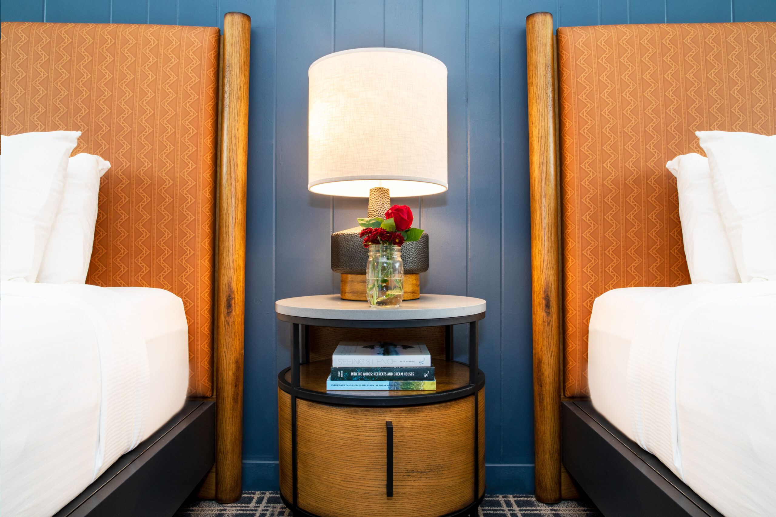 Adjacent to cozy beds, a side table accommodates a lamp, flower vase, and notebooks.