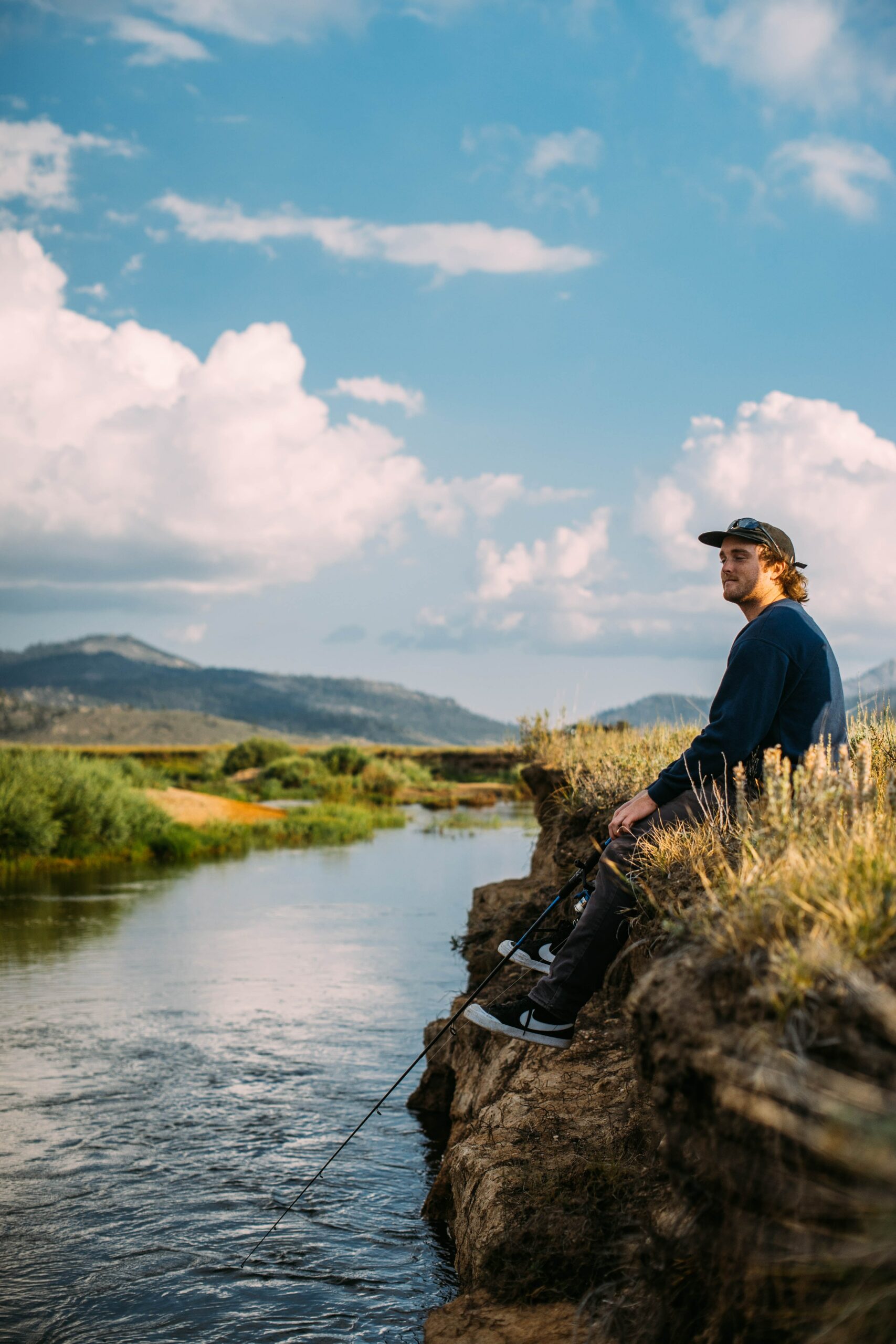 A man sits by the river's edge with a fishing rod, savoring the beauty of nature.