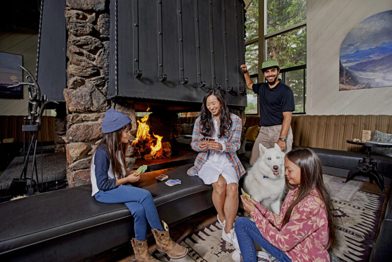 Three girls play cards by the fireplace, a smiling man stands nearby with his dog.