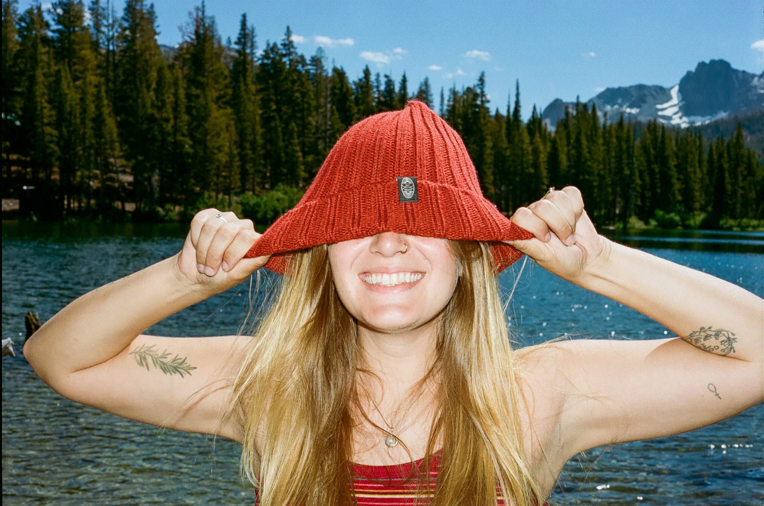 A lady in a red cap posing for a photo by the sea with tall trees in the background.