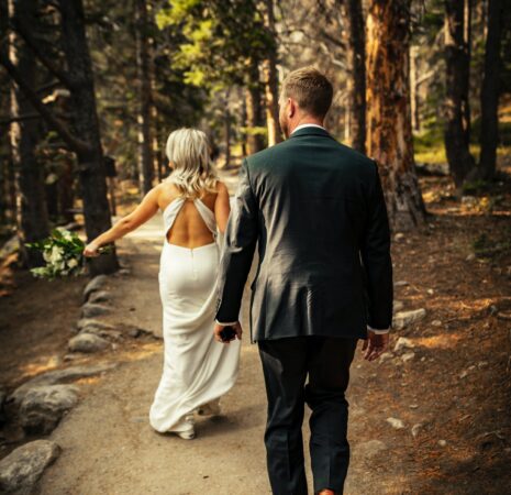 A man donning a black suit and a lady in a white gown wander through the forest, appreciating nature's beauty.