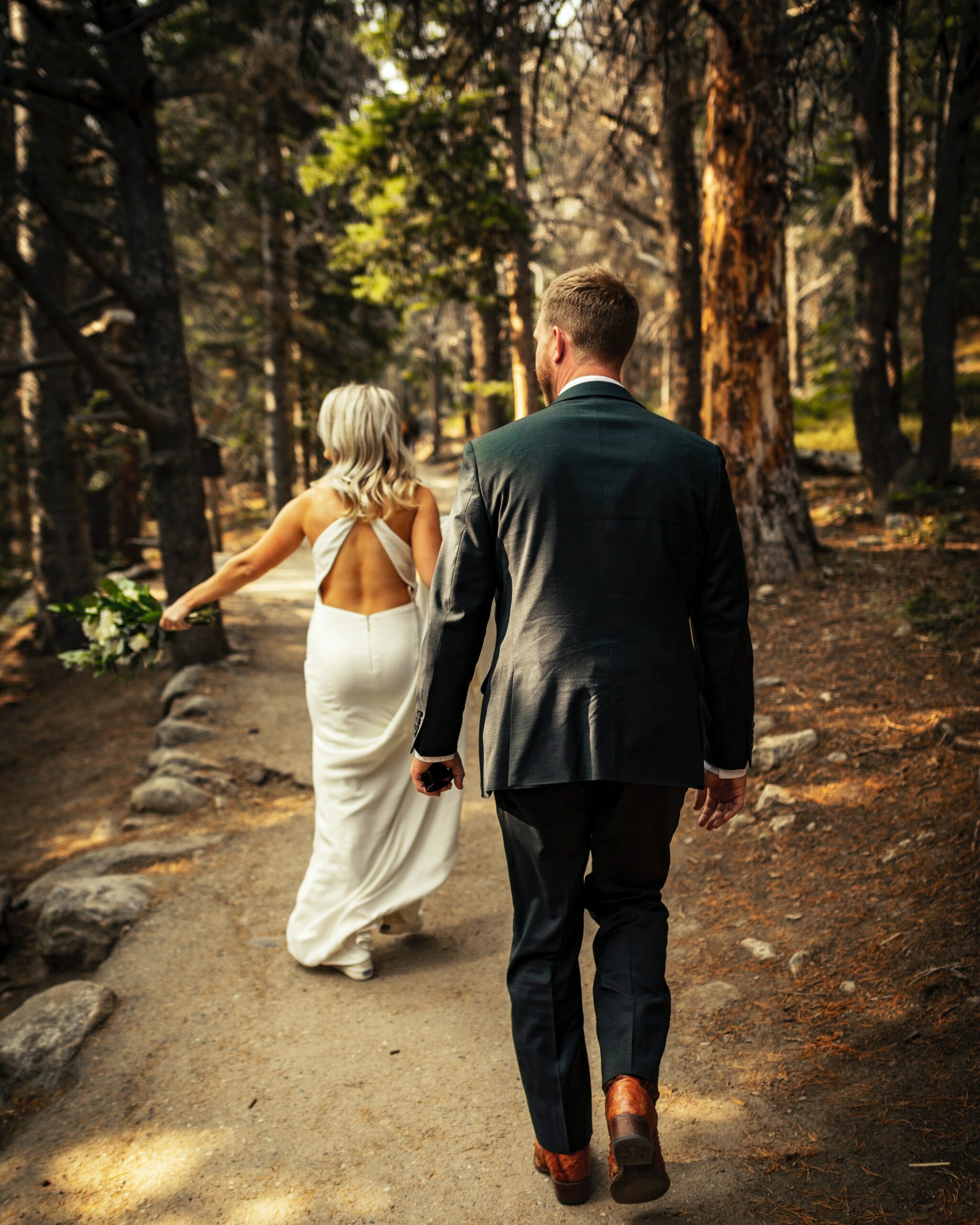 A man donning a black suit and a lady in a white gown wander through the forest, appreciating nature's beauty.