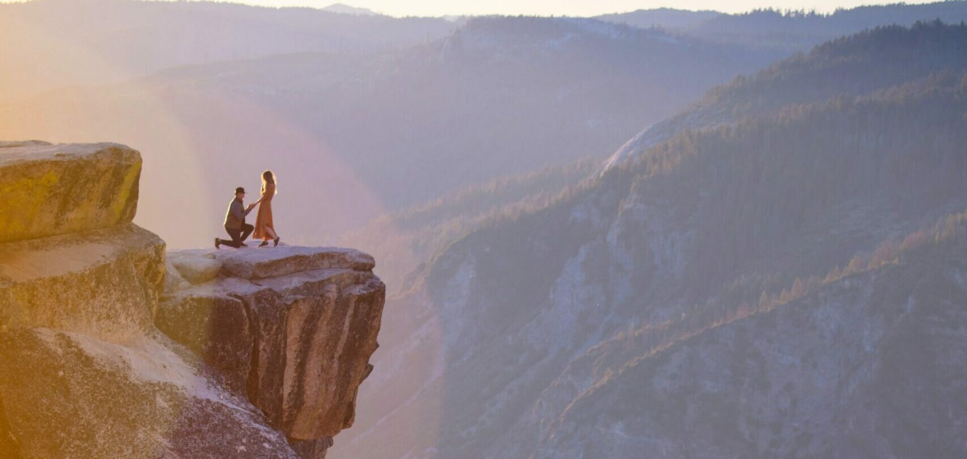 A man proposes to his partner at the mountain's edge, framed by breathtaking scenery.