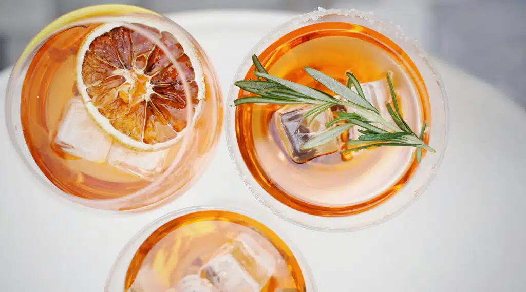Three glasses of orange-colored drinks with ice, garnished with herbs and fruit.