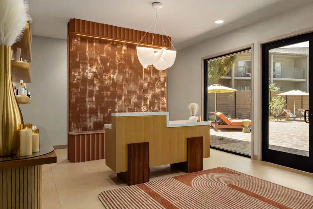 A hotel lobby with a reception desk, a glass door leading to an outdoor seating area, and a modern lighting fixture.