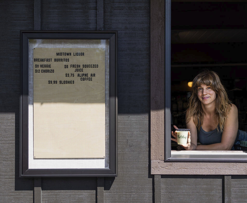 At the counter window, a woman holding hot coffee awaits customers, near a mounted menu board.
