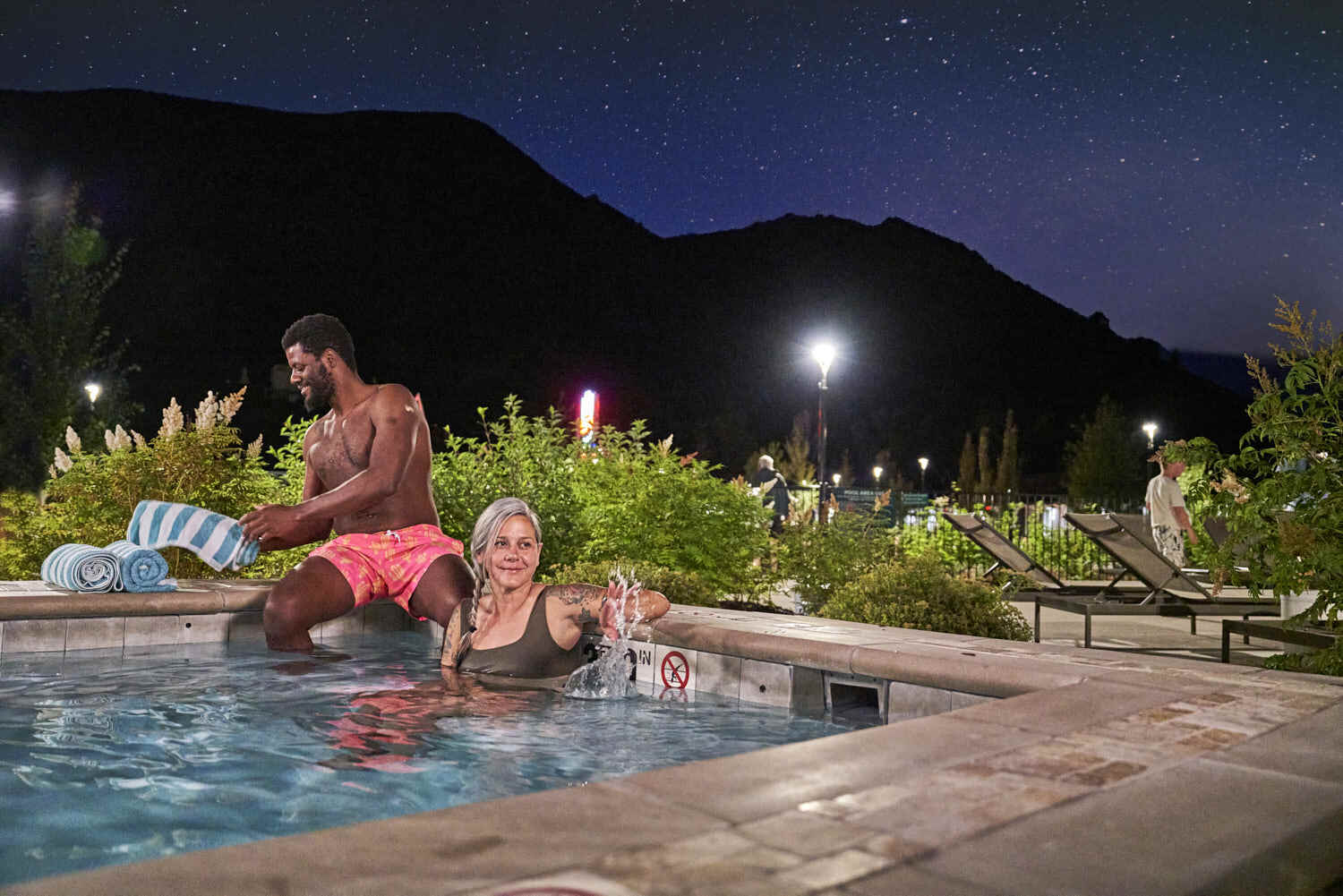 Man and woman delight in a pool bath with a scenic mountain view under a sparkling sky.