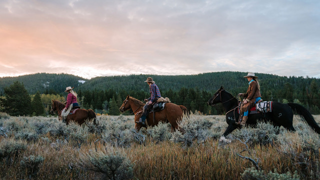 Three girls ride horses through barren land, enjoying a scenic view of green trees on mountains.