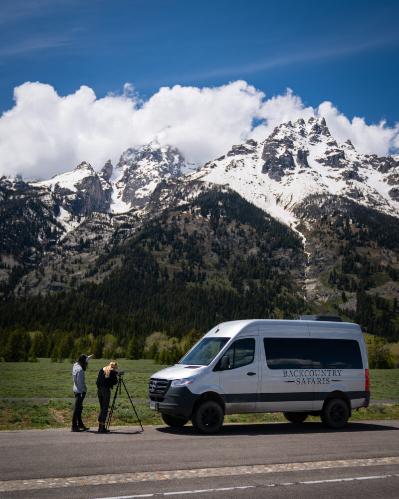 Two men capturing a photo of a snowy mountain vista with clouds, by a van labeled 