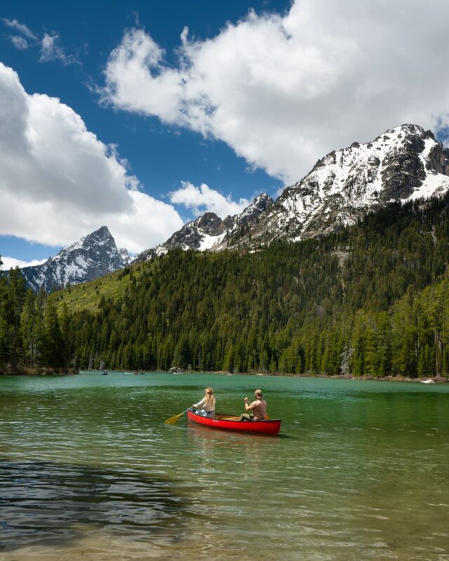 A guy and a girl sail a red boat, enjoying the scenic view of green trees and snowy mountain peaks.
