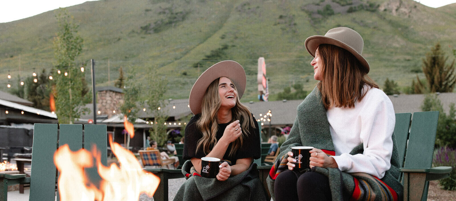 Two girls share coffee, smiles, and conversation by the bonfire, creating a cozy atmosphere.