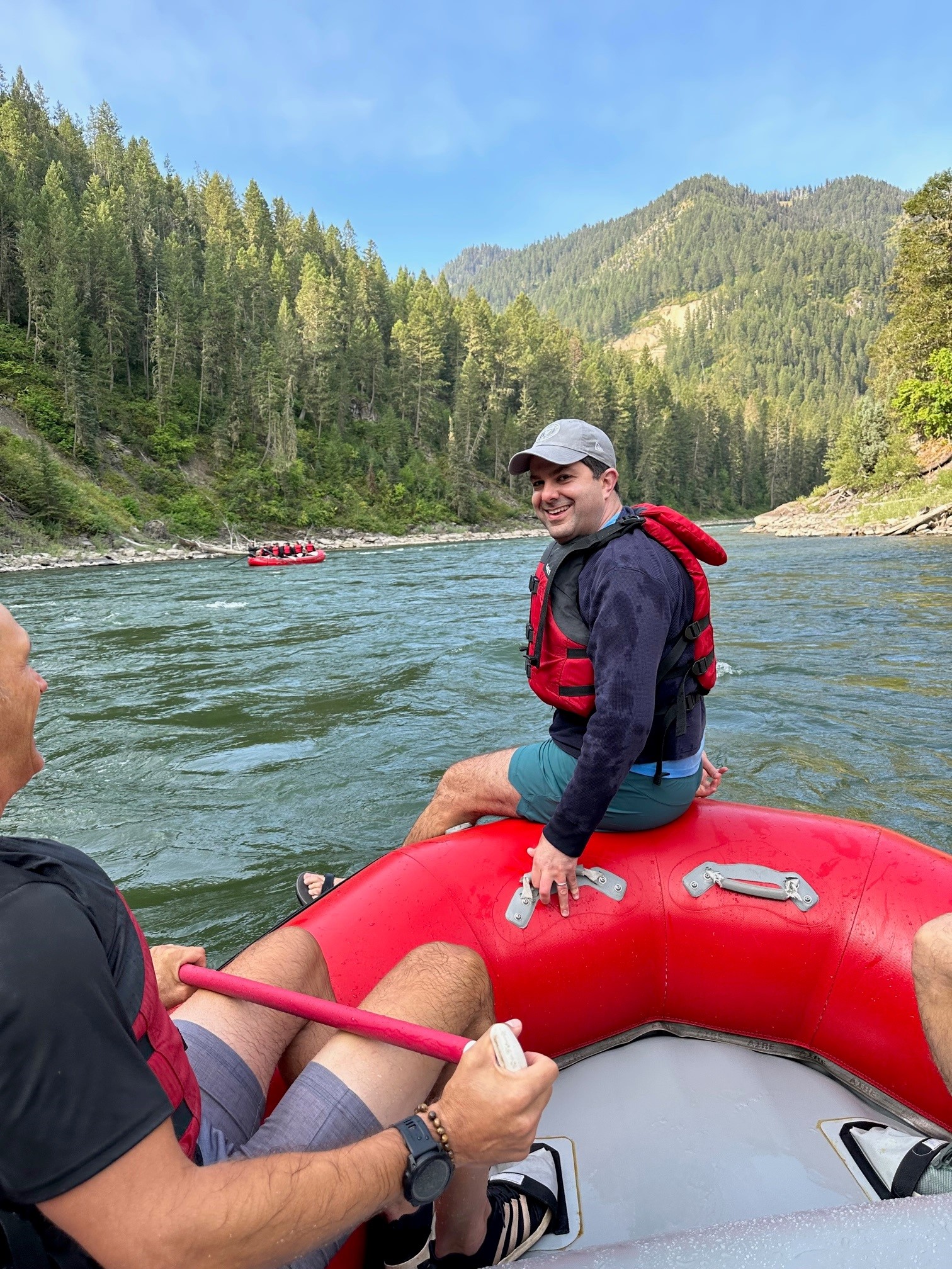 Man sits on the river rafting boat, navigating through scenic views of a mountain with tall trees.