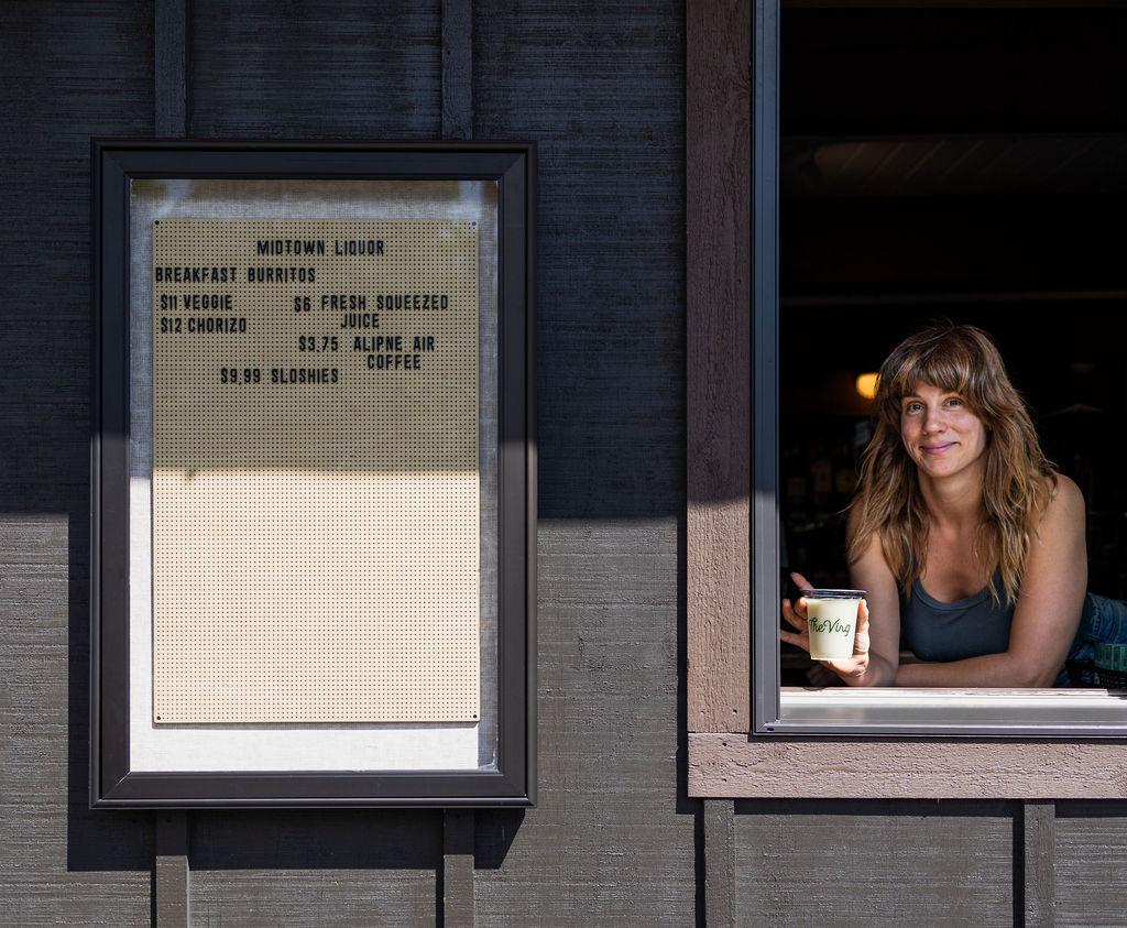 A woman at the counter window, holding a hot coffee, waits for customers, with a mounted menu board nearby.
