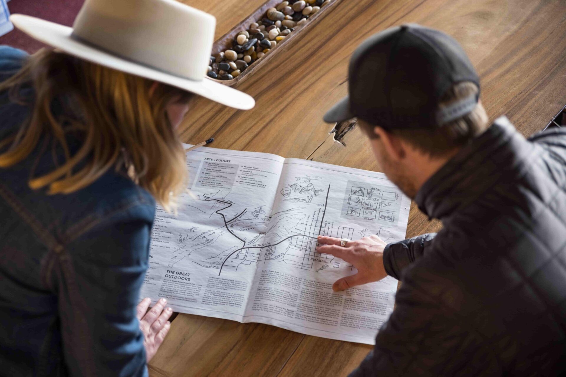 The guy in a black jacket explains a paper map to a girl in a denim jacket and hat.