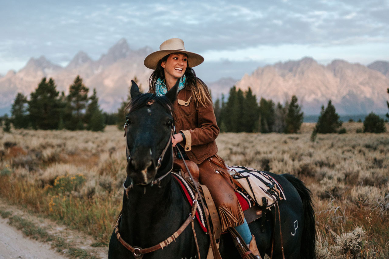 Cowgirl on horseback enjoys a stunning view of tall trees and a sparkling mountain peak.