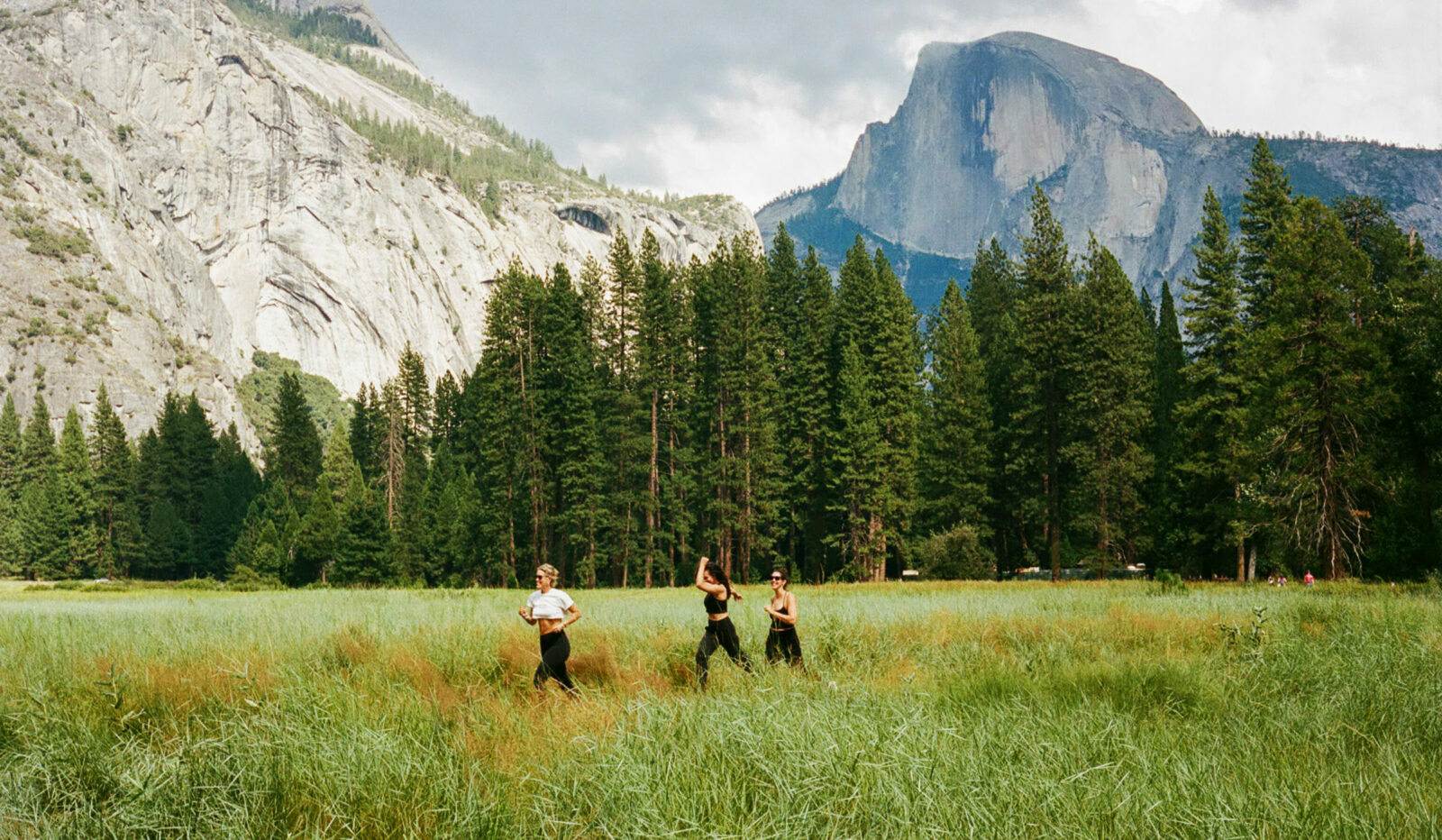 Three girls run in a grassy field, enjoying a scenic view of tall trees and mountains.