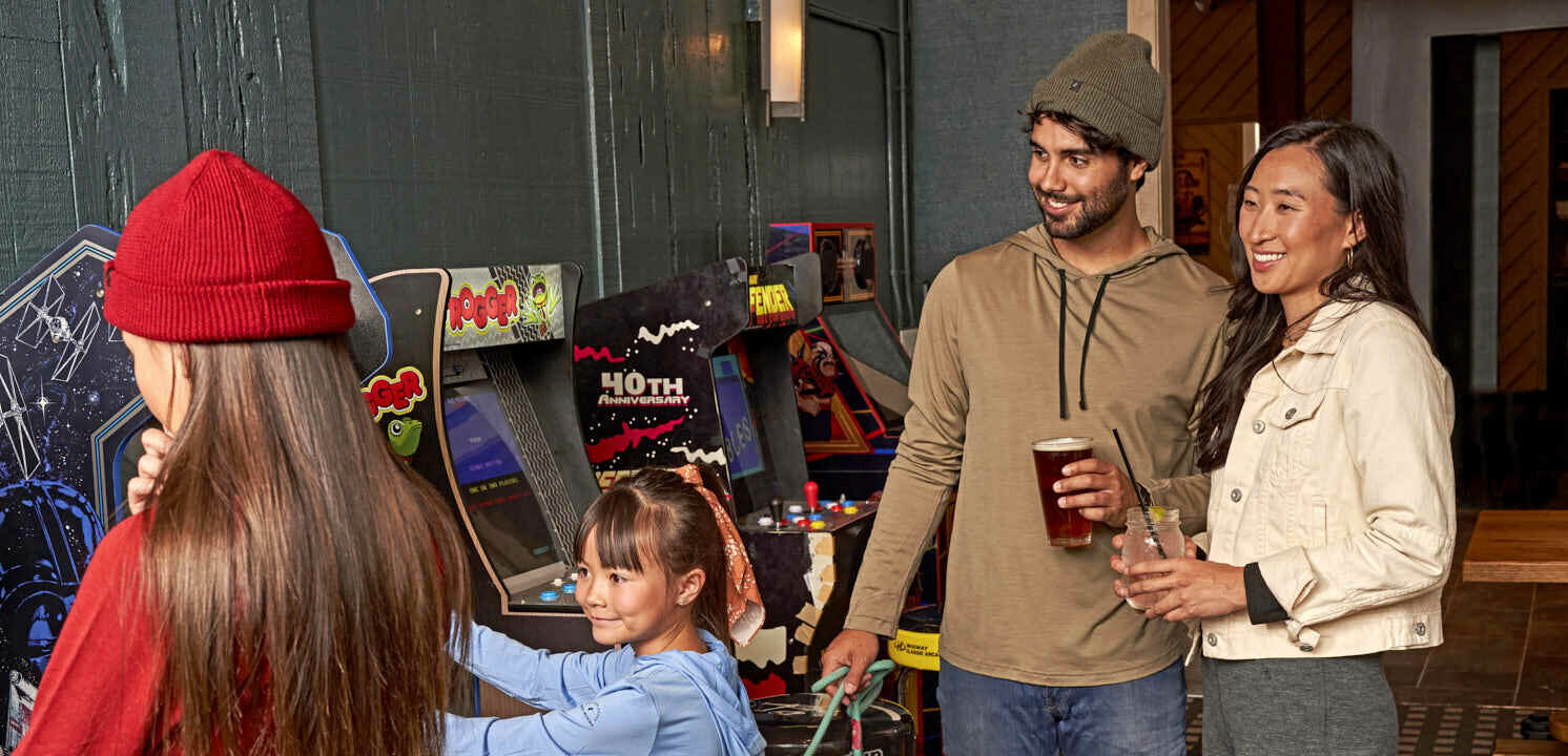 Kids play games in the corner as parents watch, holding drinks in hand.
