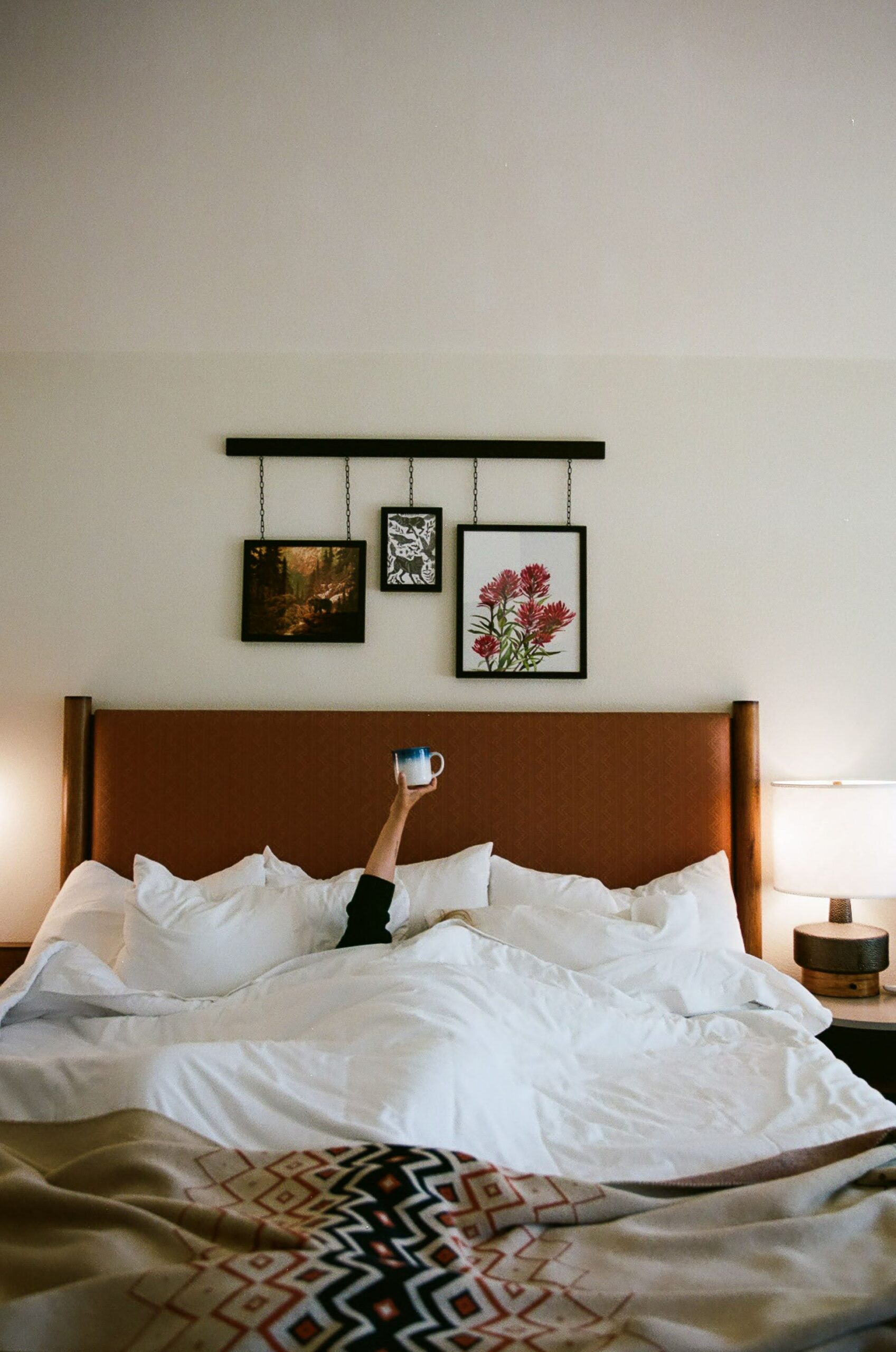 In bed, a guy raises a hand holding a coffee cup, with portraits mounted nearby.